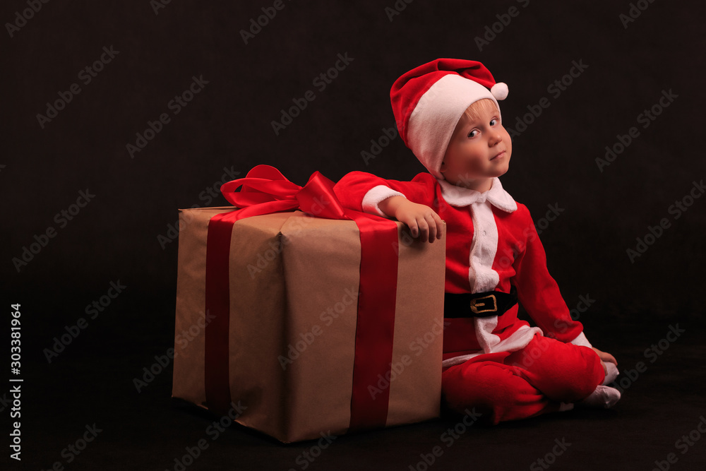 A child in a Santa Claus costume is sitting next to a gift tied with a red ribbon with a bow. A child in a Santa Claus costume is sitting next to a gift tied with a red ribbon with a bow.