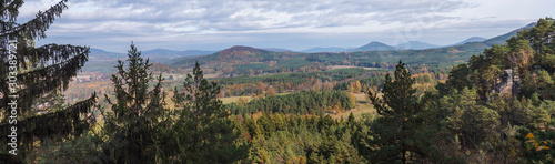 Panoramic view from Hrabencina vyhlidka on village Sloup v cechach in luzicke hory, Lusatian Mountains with autumn colored deciduous and coniferous tree forest and green hills, blue sky, white clouds