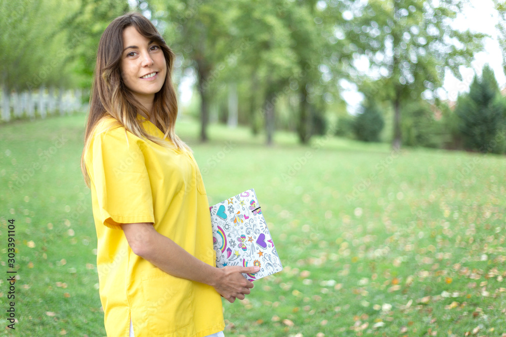 Mujer enfermera joven y sonriente vestida con uniforme amarillo sujetando una carpeta con corazones en entorno natural Mujer enfermera joven y sonriente vestida con uniforme amarillo sujetando una carpeta con corazones en entorno natural