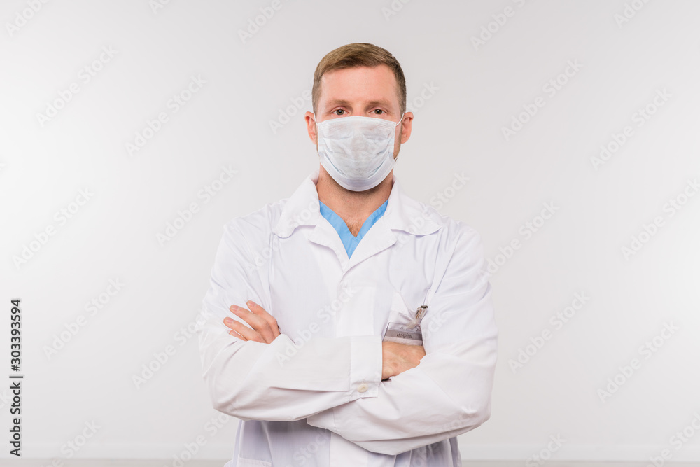 Young doctor or surgeon in whitecoat and protective mask crossing arms on chest Young doctor or surgeon in whitecoat and protective mask crossing arms on chest