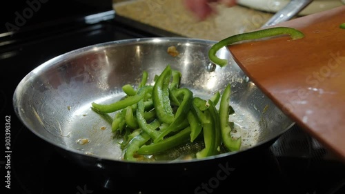 Female chef sauteing fresh sliced green peppers in frying pan on stove top oven. Bell peppers cooking in silver saute skillet.