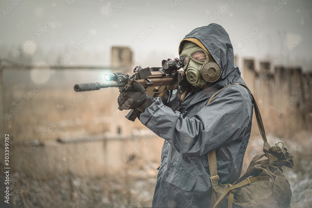 Soldier with rifle in raincoat and gas mask is walking outdoor.