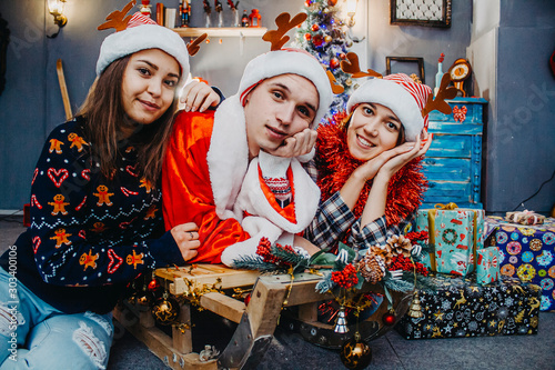 Three friends with gifts are sitting on a sleigh under a Christmas tree.