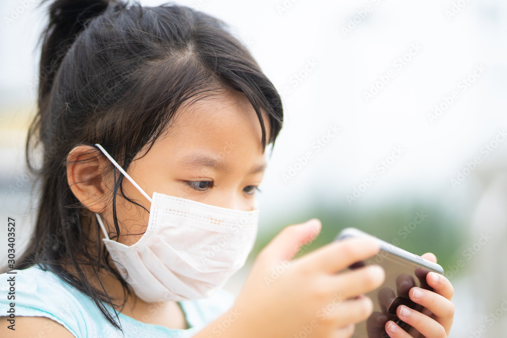 Little asian girl wearing a protective mask and holding smartphone watching cartoon with wifi and video streaming at home.sick child and Technology concept.