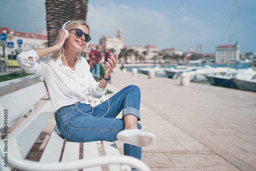 Enjoying the sound. Happy young woman with earphones listening music on smartphone while sitting on city embankment sea promenade.