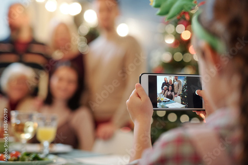Little girl with smartphone taking photo of big happy family gathered for dinner