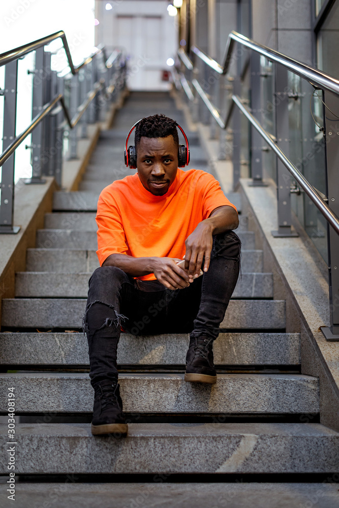 Φωτογραφία young african-american man in stylish orange hoodie sweatshirt in wireless headphones listening music and sitting on stairs