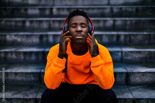 african-american man in stylish orange hoodie sweatshirt in wireless headphones listening music and enjoying music on background of stairs