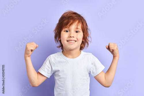 Cute caucasian kid wearing white t-shirt shows how he is strong and powerful, idolated over purple background
