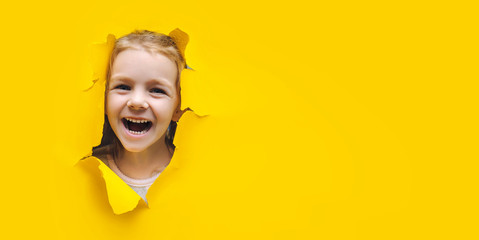 The little girl smiles happily and looks up through the hole in the yellow paper. The concept of joy, good mood and expectation of surprise. Black friday, seasonal sale. Copy space, torn paper effect.
