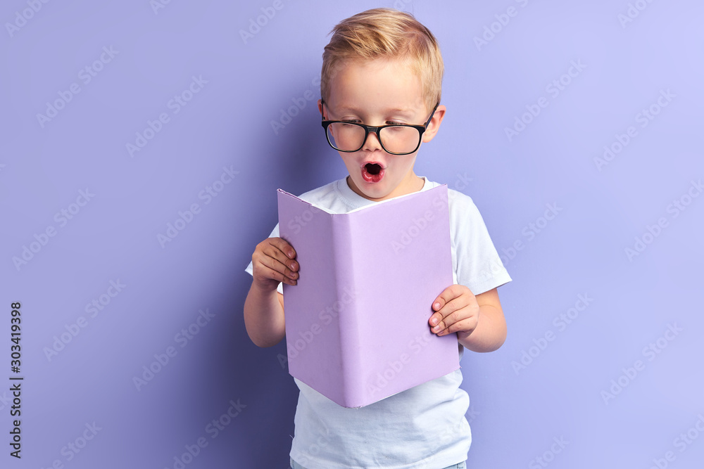 Φωτογραφία Sweet boy wearing white t-shirt and glasses for eyes reading book with purple cover, stand in shock after reading, bright emotions of kid boy