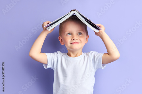 Portrait of smiling kid holding book above head, stand isolated over purple background studio. Funny education concept