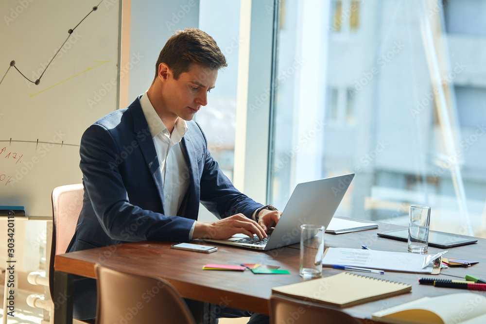 Caucasian businessman wearing a smart suit typing on laptop enthusiastically at the office space. Supplies scattered around him. Smartphone next to him. Focused and working hard. Caucasian businessman wearing a smart suit typing on laptop enthusiastically at the office space. Supplies scattered around him. Smartphone next to him. Focused and working hard.