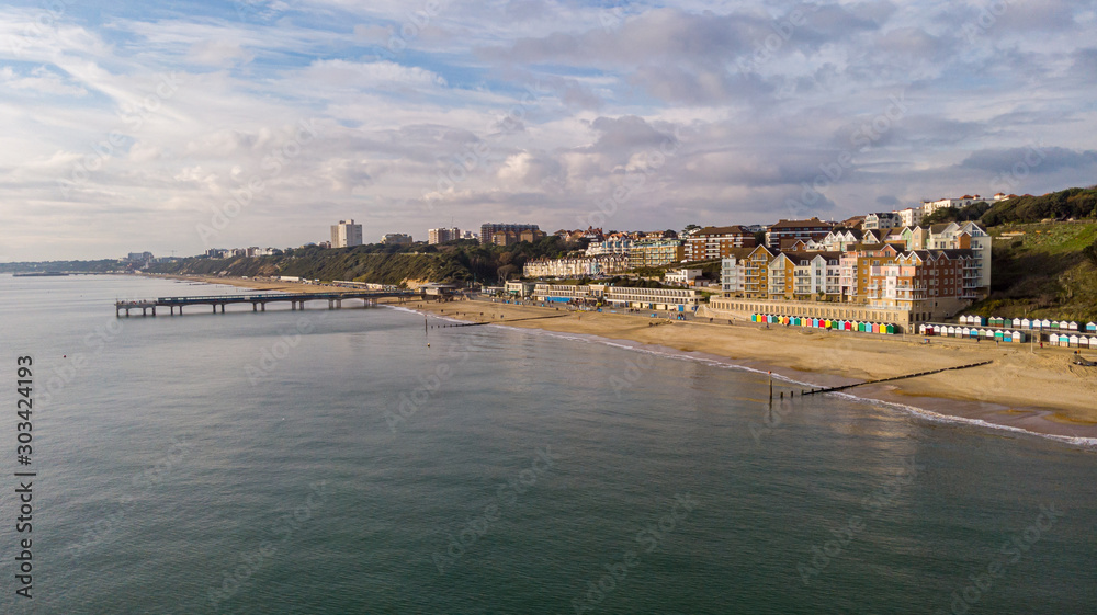 An aerial view of the Boscombe Beach with sandy beach, calm flat water, pier and building in the background under a cloudy sky with some blue sky
