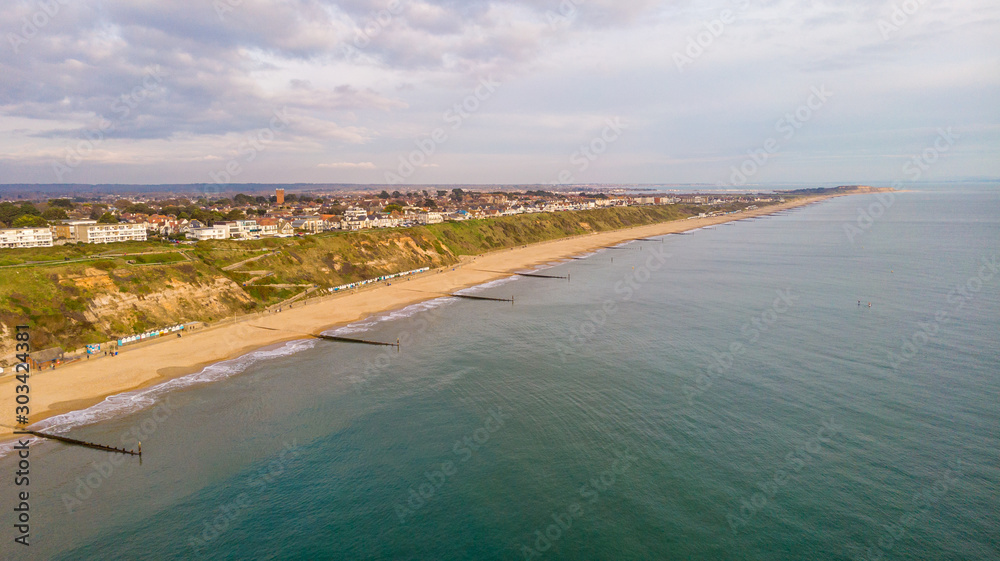 An aerial view of the Boscombe Beach with sandy beach, calm flat water, groynes (breakwaters), grassy cliff and building in the background under a cloudy sky with some blue sky