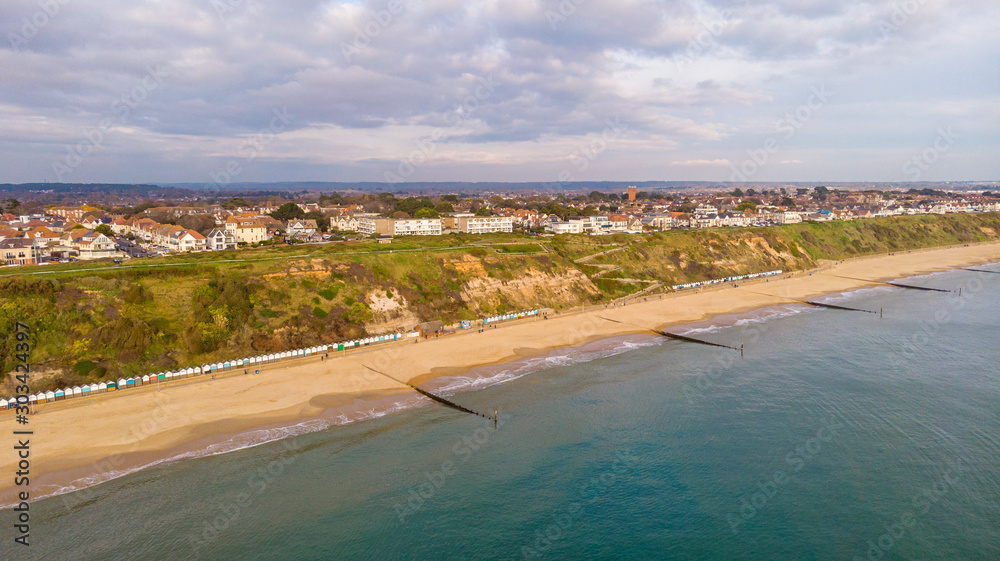 An aerial view of the Boscombe Beach with sandy beach, calm flat water, groynes (breakwaters), grassy cliff and building in the background under a cloudy sky with some blue sky