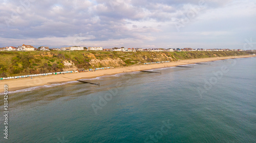 An aerial view of the Boscombe Beach with sandy beach, calm flat water, groynes (breakwaters), grassy cliff and building in the background under a cloudy sky with some blue sky