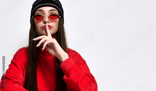Hipster girl in a fashionable black hat headdress with long hair, showing hush, shhh, secrecy, silent sign, isolated on light studio wall