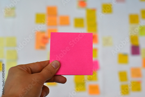 A hand is holding a pink blank sticker and there is a Kanban board of agile methodology on the background, which is a developing trend in Information Technology (IT) business.
