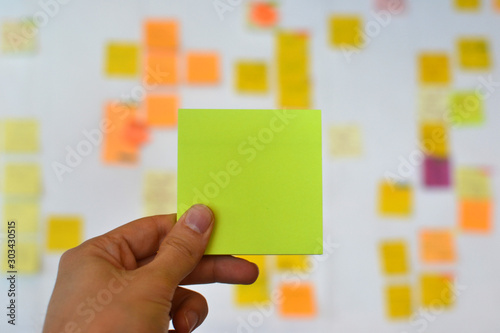 A hand is holding a yellow blank sticker and there is a Kanban board of agile methodology on the background, which is a developing trend in Information Technology (IT) business.