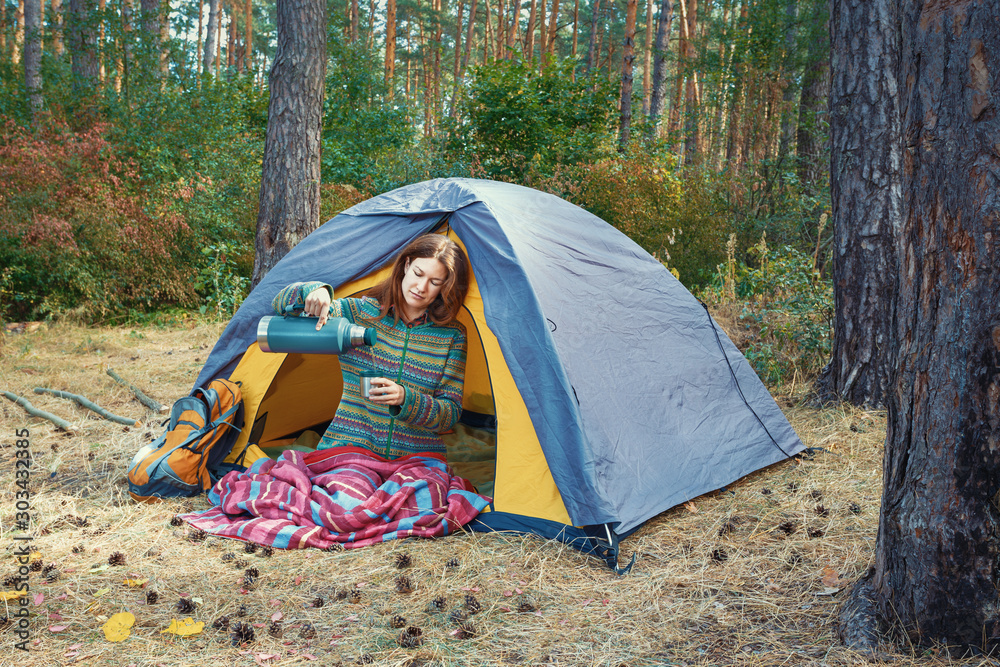 Young redhead girl pouring tea in cup from thermos, sitting in grey tent in autumn forest. Hiking, travel, weekend trip concept. Healthy active lifestyle