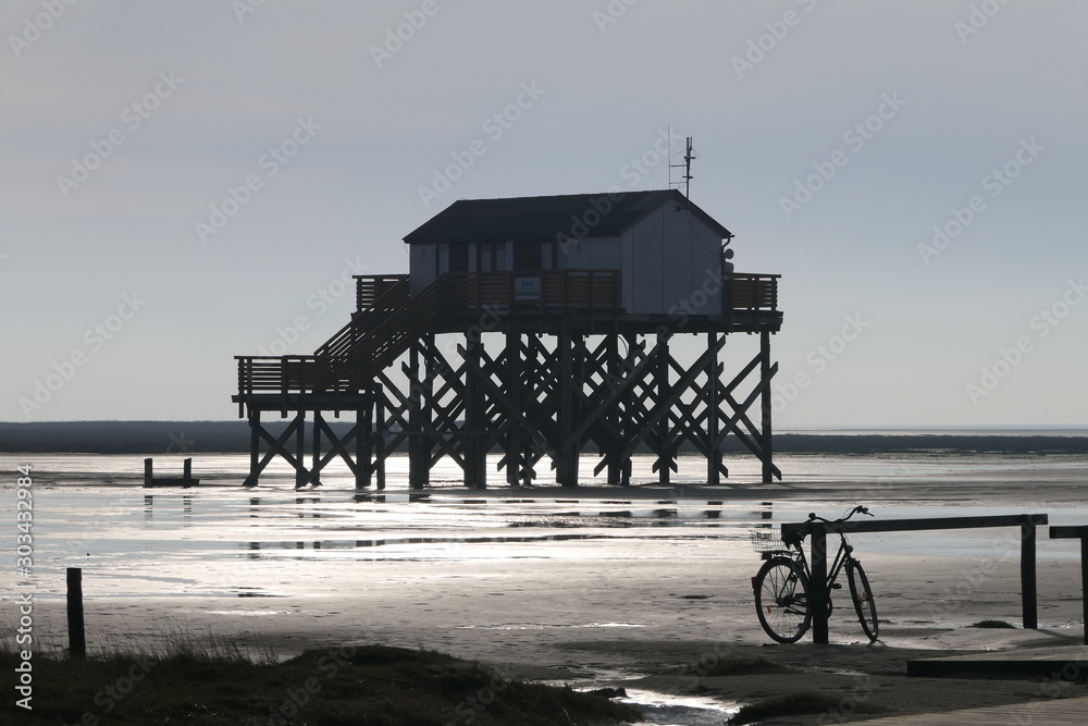 Fly at higher game. A stilt house with a staircase in cloudless sky and sunshine on the sea. At the pier is a lonely bike waiting for its owner. It's just low tide and absolute silence.