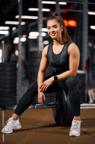 Attractive cheerful powerlifter posing in gym, sitting on barbell, looking straight at camera with broad smile, smiling joyfully, spending time in fitness club, portrait, side shot