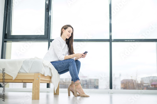 A young woman in a white shirt is talking on the phone near a large window in a hotel. Beautiful girl is negotiating while sitting on the bed. Work from home.