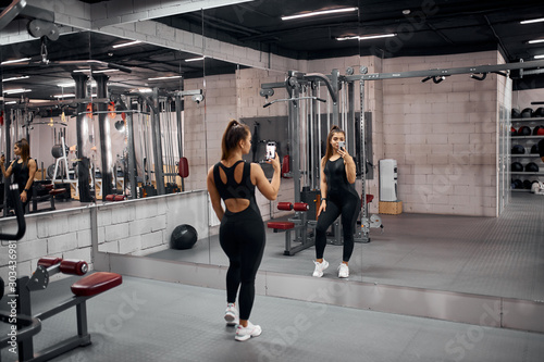 Attractive cute fitness trainer wearing black sexy leggins and short t shirt, holding phone, taking selfie, standing with back towards camera, on background of mirror wall, in gym studio