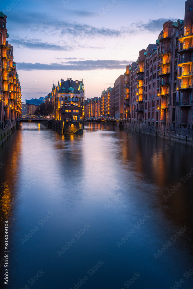 Hamburg city old port during blue hour, Germany, Europe. Historical famous warehouse district artificial illuminated. Water castle palace mirrored on river surface. Vertical orientation