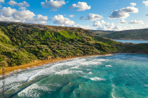 Aerial view of Ghajn Tuffieha beach and surfers in the sea. Sunset, blue sky, green mountain, waves in the sea. Winter. Malta