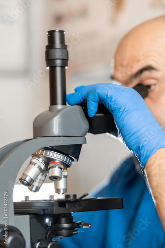 Male laboratory assistant examining biomaterial samples in a microscope. Cllose up hands in blue rubber gloves adjust microscope photo