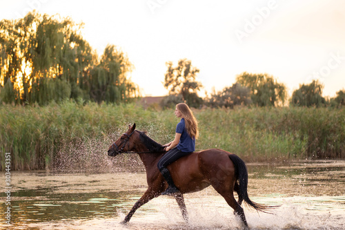 A young girl riding a horse on a shallow lake. A horse runs on water at sunset. Care and walk with the horse. Strength and Beauty