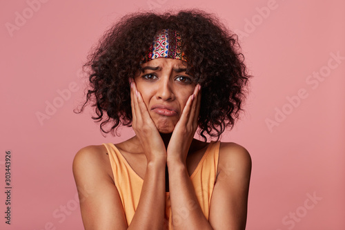 Unhappy brown-eyed young curly brunette female with dark skin looking sadly at camera and twisting her mouth, keeping palms on her cheeks while standing against pink background