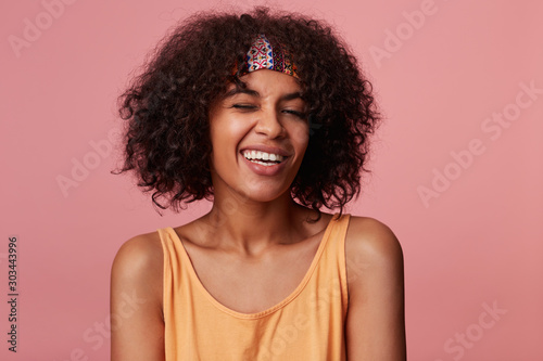Close-up of positive attractive young dark skinned lady with curly brown hair wearing casual hairstyle, looking cheerfully to camera with charming smile and giving wink, isolated over pink background