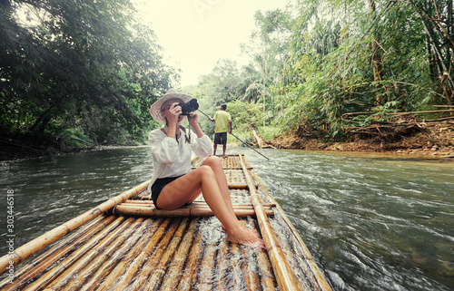 Traveling by Thailand. Pretty young woman taking photo sailing jungle river on traditional bamboo raft.