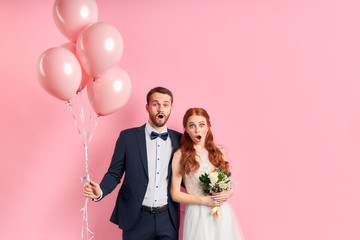 wedding couple posing together isolated over pink background. man in tuxedo holding pink air ballons, woman in white wedding dress bouquet. Woman with auburn hair, couple with opened mouth