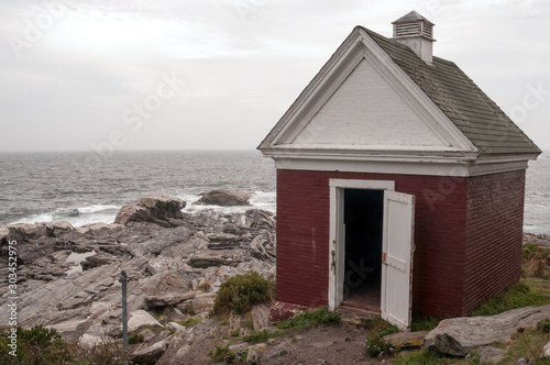 Oil shed, used for storing lighthouse oil, sitting on the rocks with the ocean surf in the background. A foggy day at Pemaquid Point Lighthouse, located in Bristol, Lincoln County, Maine