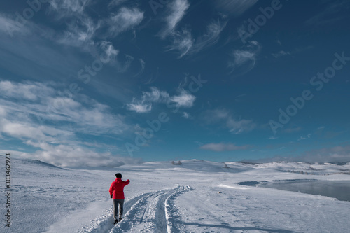 A middle age woman pointing to snowy roads in bright sportive clothes; winter natural background with snow fields, trees in frost, frozen lake, mountains; active healthy lifestyle; trekking path; Ural