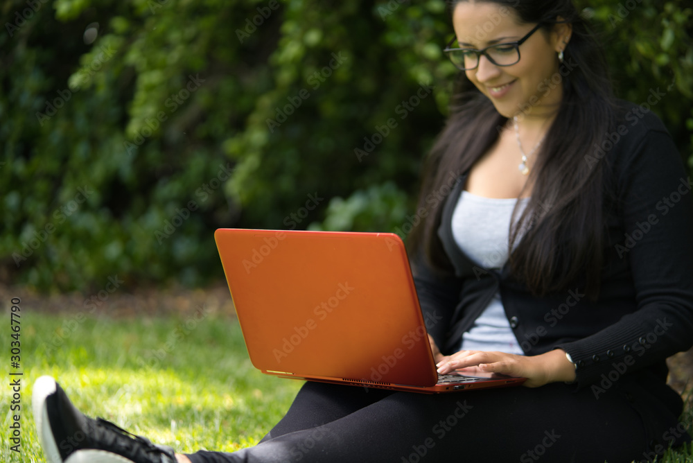 A brunette young woman wearing glasses smiling and studying with her laptop in the park. Educational concept. Natural Environment A brunette young woman wearing glasses smiling and studying with her laptop in the park. Educational concept. Natural Environment