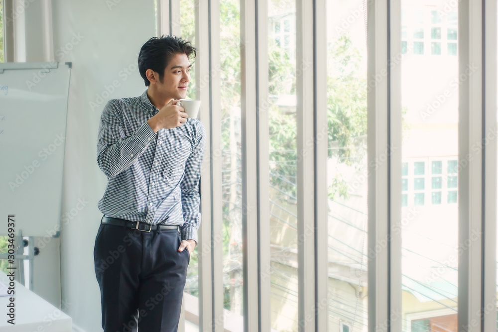 Smiling Asian businessman looking out the window while having coffee at his office. For corporate, business, finance, management, communication and technology concept. Smiling Asian businessman looking out the window while having coffee at his office. For corporate, business, finance, management, communication and technology concept.