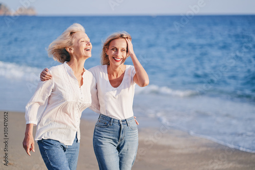 Outdoor portrait of smiling happy caucasian senior mother with her adult daughter hugging and walking on sea beach. © luengo_ua