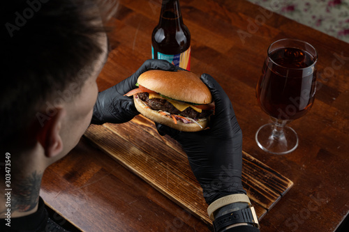 Man is black gloves is eating hamburger with beer with wooden table on the background