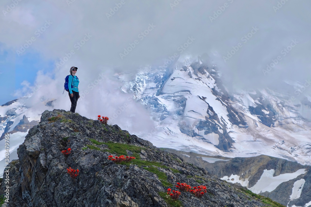 Middle age woman in sport clothes with a backpack standing on hill with red flowers by a mountain covered with glaciers and snow in clouds. Indian Paintbrush on rocks near Mt Baker. WA. USA Middle age woman in sport clothes with a backpack standing on hill with red flowers by a mountain covered with glaciers and snow in clouds. Indian Paintbrush on rocks near Mt Baker. WA. USA