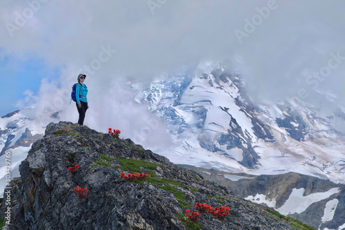 Middle age woman in sport clothes with a backpack standing on hill with red flowers by a mountain covered with glaciers and snow in clouds. Indian Paintbrush on rocks near Mt Baker. WA. USA 