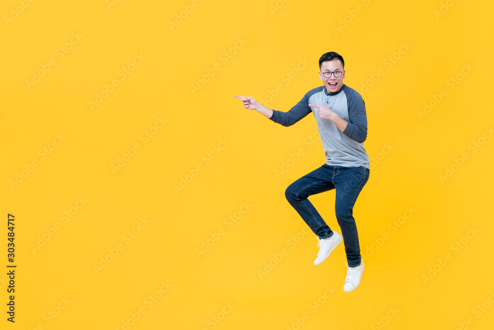Casual smiling Asian man jumping and pointing fingers to copy space aside
