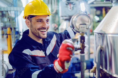 Hardworking smiling blue collar worker in protective working suit and with helmet adjusting temperature on boiler while standing in factory and holding lamp.