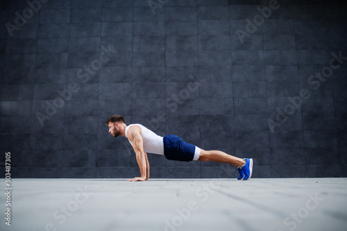 Side view of handsome Caucasian muscular bearded man in shorts and t-shirt doing push-ups. In background is gray wall.