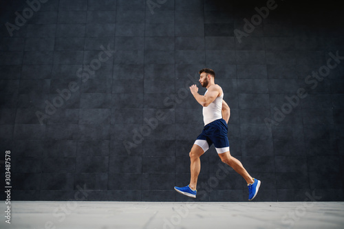 Side view of handsome caucasian muscular fit man running fast outdoors. In background is gray wall.