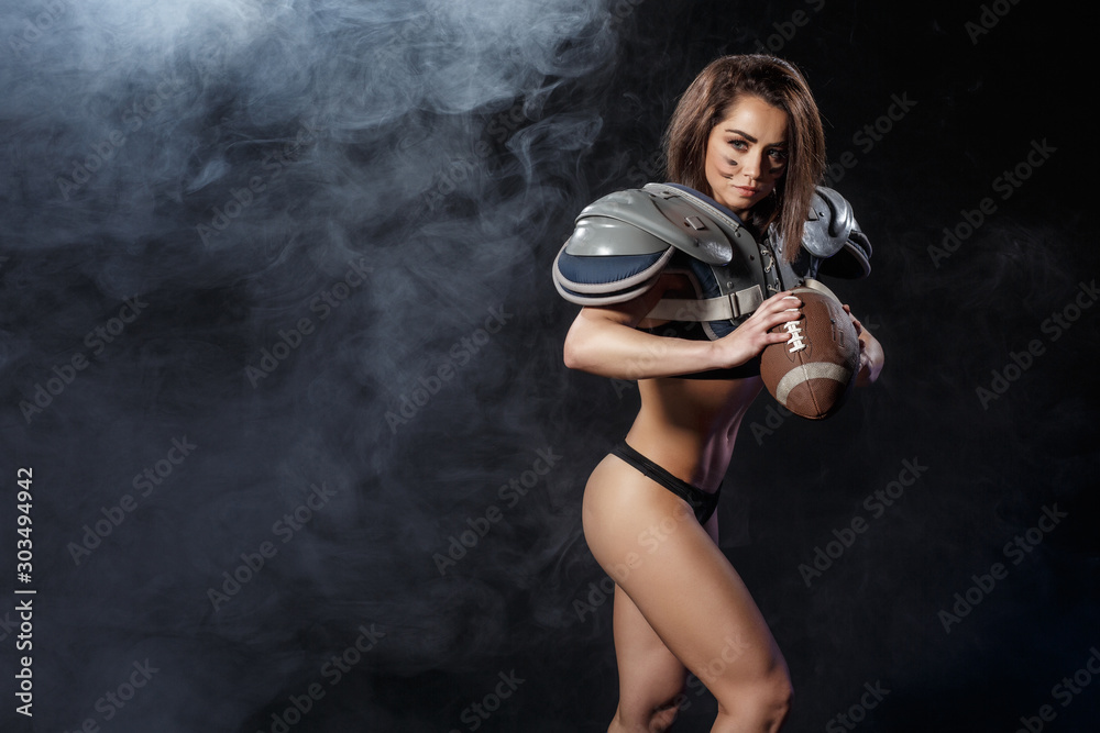 american football woman player with ball on dark smoke  background. athletic brunette fitness sexy girl as rugby player